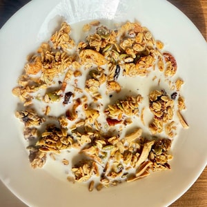 May include: A white plate filled with granola and milk. The granola is a mix of golden-brown clusters, nuts, seeds, and dried fruit. The milk is white and covers the granola. The plate is on a wooden surface.