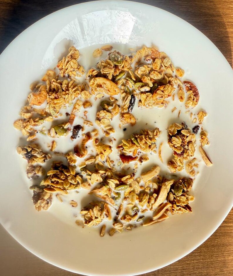May include: A white plate filled with granola and milk. The granola is a mix of golden-brown clusters, nuts, seeds, and dried fruit. The milk is white and covers the granola. The plate is on a wooden surface.