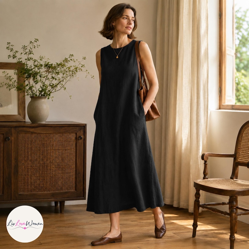 May include: A sleeveless, black midi dress with pockets. The dress is made of a textured fabric and falls to mid-calf. The model is wearing brown flats and a brown shoulder bag. The dress is simple and elegant, suitable for various occasions.