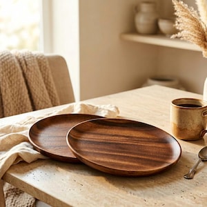 May include: Two oval wooden plates with a rich, dark brown grain sit on a light-colored table. A ceramic mug with coffee and a small spoon are also on the table. The scene is bathed in natural light, suggesting a dining or kitchen setting.