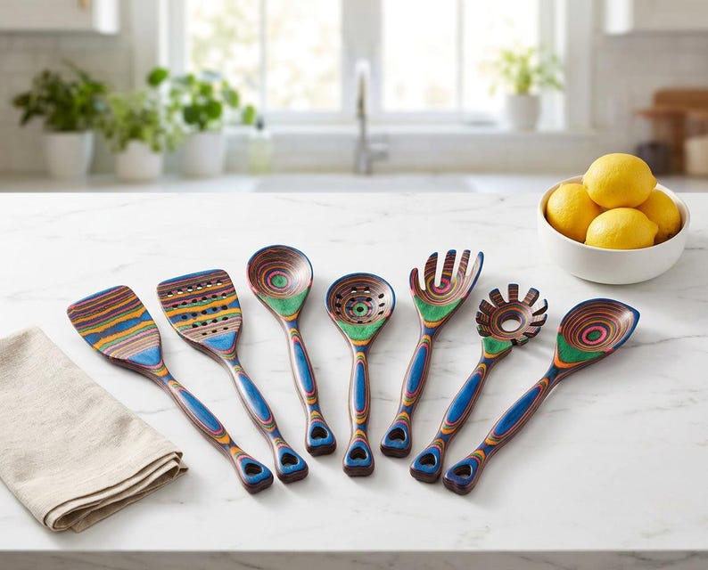 Puede incluir: Un juego de siete utensilios de cocina de madera coloridos, que incluyen una esp&aacute;tula, una cuchara ranurada y una cuchara para servir. Los utensilios tienen mangos azules y est&aacute;n colocados sobre una encimera blanca junto a un pa&ntilde;o de lino y un cuenco de limones.