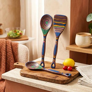 Puede incluir: Un juego de tres utensilios de cocina de madera con dise&ntilde;os coloridos de c&iacute;rculos conc&eacute;ntricos. El juego incluye una cuchara, una esp&aacute;tula y una cuchara m&aacute;s peque&ntilde;a, todos sobre una tabla de cortar de madera. Tambi&eacute;n hay un lim&oacute;n y dos tomates cherry.