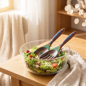 Puede incluir: Un cuenco de ensalada de vidrio transparente lleno de verduras frescas, tomates y almendras. Dos utensilios de ensalada de madera con mangos a rayas azules, verdes y marrones descansan en el cuenco. El cuenco est&aacute; sobre una mesa de madera.