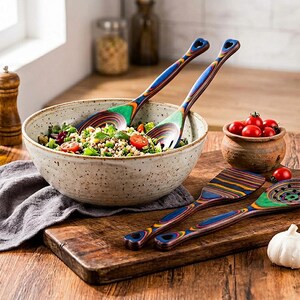 Puede incluir: Un cuenco de cer&aacute;mica moteada lleno de ensalada, acompa&ntilde;ado de coloridos utensilios y cubiertos de madera para ensalada. Los utensilios de madera tienen un dise&ntilde;o de arco&iacute;ris y est&aacute;n sobre una tabla de cortar de madera. Tambi&eacute;n hay una peque&ntilde;a olla de tomates y un diente de ajo.