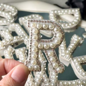 Könnte beinhalten: Nahaufnahme des Buchstabens "R", verziert mit Kunstperlen und kleinen, klaren Strasssteinen. Der weiße Buchstabe hat einen silbernen Rand und gehört zu einem Satz dekorativer Buchstaben. Weitere Buchstaben sind im Hintergrund zu sehen.