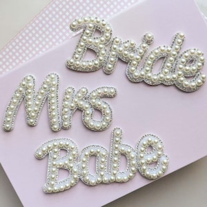May include: Three decorative words: "Bride", "Mrs", and "Babe". Each word is made with white faux pearls and outlined with silver rhinestones. The words are arranged on a light pink surface, ideal for bridal decorations.
