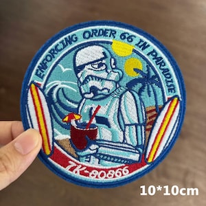 Könnte beinhalten: Runder, bestickter Aufnäher in Blau und Weiß mit einer Cartoon-Figur, die einen Cocktail trinkt, Surfbrettern und Palmen. Der Text lautet "ENFORCING ORDER 66 IN PARADISE". Der Aufnäher misst 10 x 10 cm.