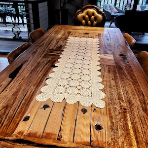 May include: A rectangular cream-coloured crochet table runner featuring a repeating circular motif and a scalloped edge. The runner is centred on a large, dark brown wooden dining table with visible wood grain and natural knots in the surface.
