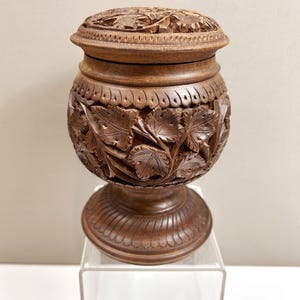 May include: A dark brown wooden lidded jar featuring intricate hand-carved leaf patterns around the body and a matching carved lid. The vessel has a rounded midsection, a flared base with decorative scalloped detailing, and a smooth finish.
