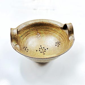 Può includere: Scolapasta in ceramica nei toni del beige e del marrone. La ciotola presenta piccoli fori di drenaggio e due manici. Questo utensile da cucina è progettato per scolare i liquidi dagli alimenti, con un diametro di circa 25 cm.