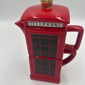 May include: A red ceramic teapot shaped like a British telephone box. The teapot features black grid panels, a handle, a spout, and a lid topped with a gold-colored crown finial. The word TELEPHONE is printed in black letters on a white background.
