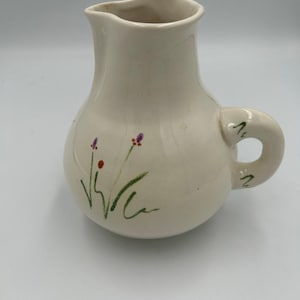 May include: A cream-colored ceramic pitcher with a rounded body and a single handle. The exterior features a hand-painted design of thin green stems with small purple and red floral accents. The handle also includes small green painted marks.