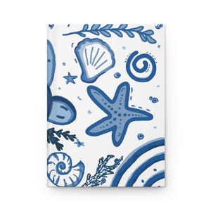 May include: A white journal with a blue watercolor design featuring seashells, starfish, and other marine elements. The cover has a clean, minimalist aesthetic with a nautical theme, perfect for writing or sketching.