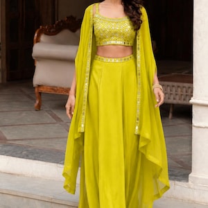 May include: A lime green three-piece ensemble consisting of a sleeveless crop top, a floor-length skirt, and a matching cape. The top and the waistband of the skirt feature intricate mirror work embroidery. The cape has a sheer, flowing design.
