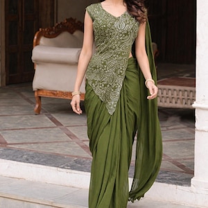 Könnte beinhalten: Ein olivgrünes Saree-Ensemble. Das Oberteil hat einen V-Ausschnitt und aufwendige silberne Stickereien. Der drapierte Saree fließt anmutig und erzeugt eine elegante Silhouette. Das Outfit ist mit goldenen Armbändern und Ohrringen ausgestattet.