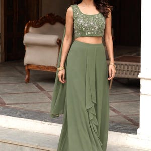 May include: An olive green two-piece ensemble consisting of a sleeveless crop top and a floor-length skirt. The top features intricate silver floral embroidery and beadwork. The skirt has a draped front panel with a ruffled edge and a matching scarf.