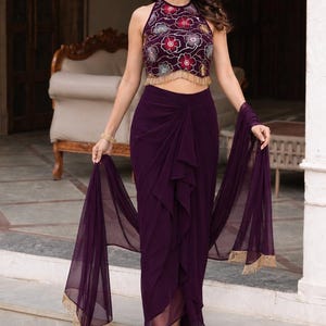 May include: A deep purple two-piece outfit consisting of a sleeveless halter-neck crop top with embroidered floral patterns and gold fringe trim, paired with a matching high-waisted draped skirt and a sheer purple shawl with gold fringe edges.