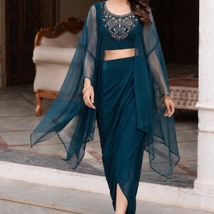 May include: A teal three-piece ensemble consisting of a sleeveless crop top with intricate silver and gold embroidery around the neckline, a matching draped dhoti-style skirt, and a sheer, lightweight, long-sleeved cape overlay in the same teal colour.
