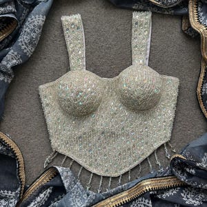 Puede incluir: Top bustier plateado con cuentas, con escote corazón y tirantes finos. La parte superior está adornada con cuentas iridiscentes y tiene un dobladillo festoneado. Una bufanda azul y gris estampada con ribete dorado enmarca la parte superior.