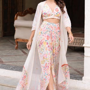 May include: A three-piece floral print outfit comprising a crop top, high-waisted skirt with a front slit, and a long, sheer, white jacket. The outfit features a colourful floral pattern with pink, yellow, and blue flowers. The model is wearing gold sandals.