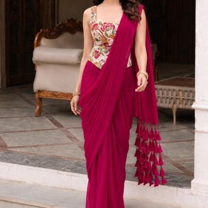 May include: A magenta saree featuring a sleeveless bodice with a cream-colored floral print. The garment includes a long, flowing skirt with draped fabric and a matching magenta pallu that hangs over the shoulder, finished with a row of decorative tassels.