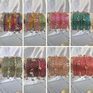 May include: A selection of colourful bangles in shades of pink, green, and red. Each bangle features small, dangling gold-coloured charms. The bangles are presented on a white surface.