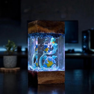 May include: A rectangular lamp featuring a dark wood base and top with a clear resin center. Inside the resin are three blue and yellow turtle-like figures, including a small one, a medium one, and a large one, surrounded by blue water-like accents.