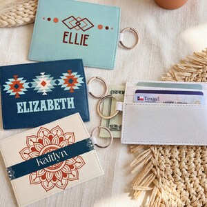 May include: A collection of personalized cardholders in various colors. The cardholders feature names like "Ellie", "Elizabeth", and "Kaitlyn", along with decorative designs. One white cardholder is open, displaying cards and cash. Each cardholder has a keyring.