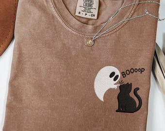May include: A brown t-shirt featuring an embroidered design of a white ghost touching the nose of a black cat with the text BOOOop. The shirt has a Comfort Colors label at the neck and is paired with a thin silver chain necklace with a small pendant.