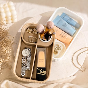 Op de afbeelding: Een beige organizer dienblad gevuld met sieraden, make-up borstels en schoonheidsproducten. Items zijn onder meer een zilveren horloge, ringen, oorbellen en een fles foundation. Een apart wit dienblad bevat huidverzorgingsproducten en een klein schaaltje met sieraden.