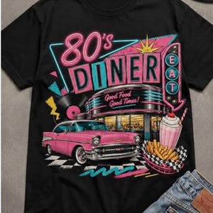 Op de afbeelding: Een zwart t-shirt met een grafische print van een roze klassieke auto, een diner-gebouw met de tekst 80's DINER en EAT, een milkshake en frietjes. Het ontwerp bevat een vinylplaat, neon-stijl accenten en de tekst Good Food Good Times.