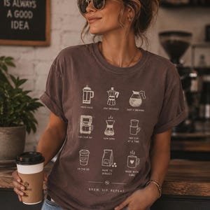 May include: A dark brown short-sleeve t-shirt featuring a white graphic print of nine different coffee brewing methods with corresponding text labels and the phrase "BREW. SIP. REPEAT." printed at the bottom of the design.