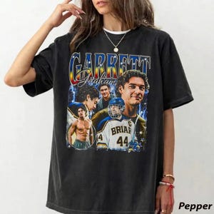 Può includere: Una t-shirt nera a maniche corte con una stampa grafica recante il testo "GARRETT Graham". Il design include diverse immagini di un uomo, una a torso nudo e una con indosso una maglia da hockey bianca con la scritta "BRIAR 44" sul davanti.