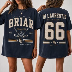 May include: A navy blue short-sleeve t-shirt featuring a Briar University Hockey Club graphic on the front with crossed hockey sticks and the text Play Hard, Stay Humble. The back displays the name Di Laurentis and the number 66 in large font.