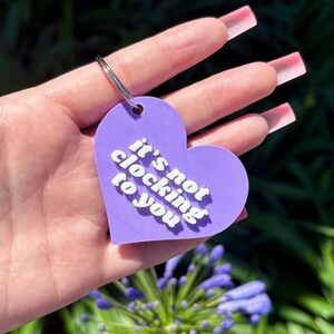 May include: A purple heart-shaped keychain with the phrase "it's not clocking to you" in white, sans-serif font. The keychain has a silver ring and is held against a background of green foliage and blue flowers.