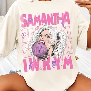 May include: An off-white short-sleeve t-shirt featuring a graphic print of a character with pigtails biting a purple twenty-sided die. The design includes the name SAMANTHA in large pink letters, smaller skulls, lightning bolts, and the text IWKYM.