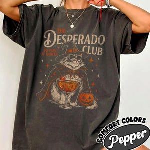 May include: A pepper-colored t-shirt featuring a graphic of a cat wearing a witch hat and sunglasses, holding a cocktail glass. The text reads The Desperado Club So Fun It Hurts. The design includes stars and a jack-o'-lantern pumpkin.