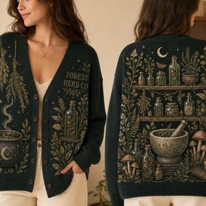 May include: A dark green V-neck button-down cardigan featuring a graphic print of a steaming cauldron on the front and shelves filled with apothecary jars, herbs, mushrooms, and a mortar and pestle on the back, with the text Forest Herb Co.