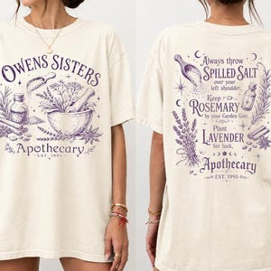 Op de afbeelding: Een ivoorkleurig t-shirt met paarse grafische prints. De voorkant toont een vijzel met stamper en kruiden en de tekst Owens Sisters Apothecary Est. 1995. De achterkant bevat botanische illustraties en tekst over zout, rozemarijn en lavendel.