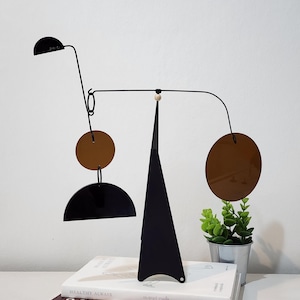 Puede incluir: Una escultura móvil moderna de metal negro y bronce con formas geométricas. La escultura presenta una base triangular alta y un alambre horizontal con elementos circulares y semicirculares. La escultura se exhibe sobre dos libros.
