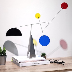 Op de afbeelding: Een moderne mobiele sculptuur met een geometrisch ontwerp. Het heeft een zilveren metalen basis en draadstructuur, die een zwarte halve cirkel, gele en blauwe cirkels en een rode cirkel ondersteunt. De sculptuur wordt tentoongesteld op een houten oppervlak met boeken en kleine planten.