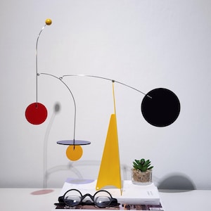 Puede incluir: Una escultura cinética con finos alambres de metal plateado, una esfera amarilla, un círculo rojo, un círculo amarillo pequeño, un círculo azul oscuro, un círculo negro grande y una base triangular amarilla sobre dos libros apilados.