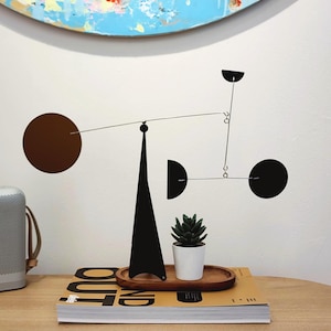May include: A contemporary black metal mobile sculpture featuring circular and geometric shapes. The sculpture is displayed on a wooden surface with a small potted succulent and a book. An abstract painting hangs in the background.