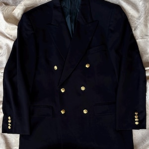 May include: A double-breasted navy blue blazer featuring six gold-toned metal buttons on the front, two flap pockets at the waist, and four smaller gold-toned buttons on each sleeve cuff. The jacket has wide lapels and a structured silhouette.