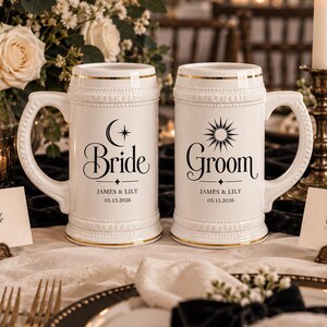 Puede incluir: Dos jarras de cerveza de cerámica blanca con bordes y asas dorados. Una presenta un icono de luna y el texto "Bride JAMES & LILY 05.15.2026", la otra un icono de sol y el texto "Groom JAMES & LILY 05.15.2026" en letras negras.