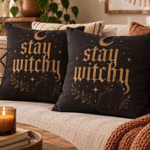 Puede incluir: Dos cojines cuadrados negros con un estampado gráfico de color dorado. El diseño incluye el texto stay witchy en una fuente gótica, una luna creciente, estrellas y un gato sentado rodeado de delicadas ilustraciones de hojas y enredaderas.