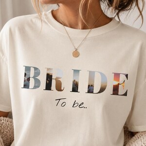 Puede incluir: Una camiseta de manga corta de color crema con la palabra BRIDE en letras mayúsculas grandes, donde cada letra contiene una escena fotográfica diferente de una pareja. Debajo de la palabra BRIDE, el texto To be.. está impreso en letra negra.