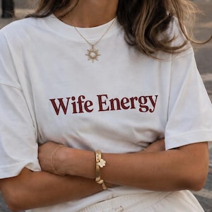 Puede incluir: Una camiseta blanca de manga corta con el texto Wife Energy impreso en una fuente serif de color rojo oscuro en el pecho, combinada con unos pantalones cortos blancos a juego con cintura elástica y cordón ajustable en el centro.