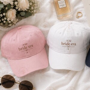 Puede incluir: Tres gorras de béisbol en rosa, blanco y azul marino, cada una bordada con el texto 'in my bride era' y un pequeño icono de lazo. Las gorras están dispuestas con un frasco de perfume, una invitación de boda, un anillo de oro, gafas de sol y un pasaporte.