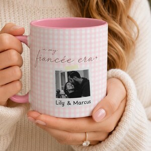 Puede incluir: Una taza de cerámica con estampado de cuadros vichy en rosa y blanco, con asa e interior de color rosa. La taza presenta el texto "in my fiancée era" en letra cursiva sobre una fotografía en blanco y negro de una pareja, con los nombres "Lily & Marcus".